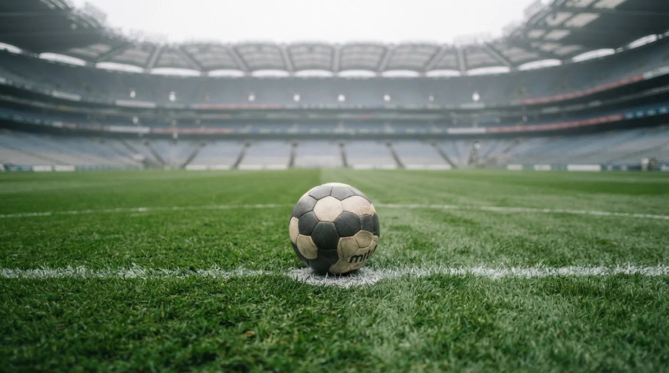 Ein Fußball liegt auf dem Elfmeterpunkt eines leeren Rasenplatzes, im Hintergrund eine Tribüne im Dunst.