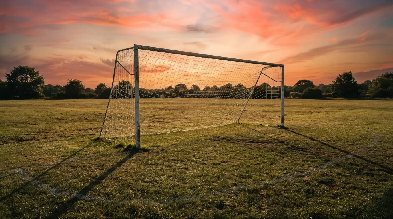 Ein Fußballtor mit Netz bei Sonnenuntergang auf einem leeren Platz, die Schatten der Torpfosten liegen lang auf dem Gras.