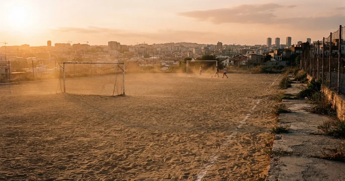 Ein sandiger Fußballplatz am Rand einer Stadt in warmem Abendlicht, Staub in der Luft und zwei Tore in der Ferne.
