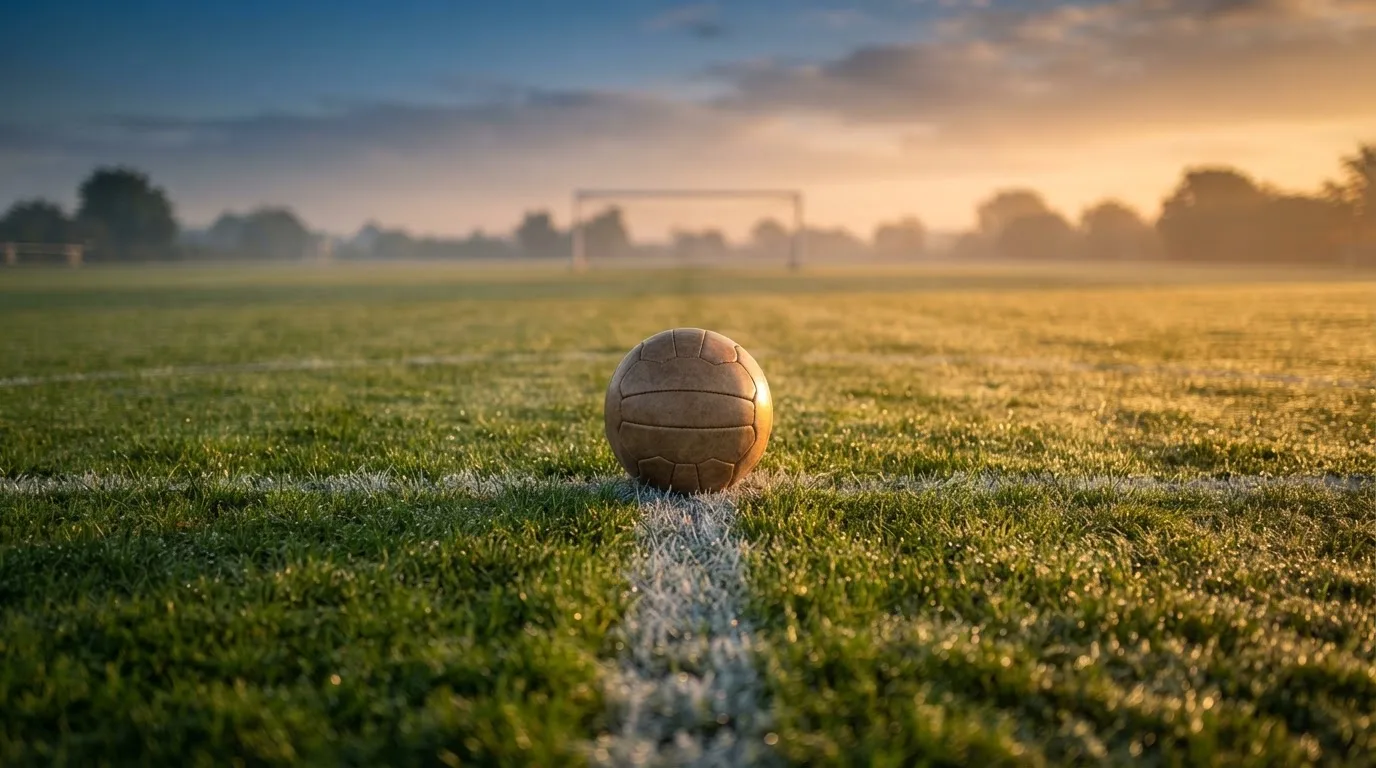 Ein Fußball liegt auf dem Anstoßpunkt eines Rasens bei Sonnenaufgang, Tau glitzert auf den Halmen.