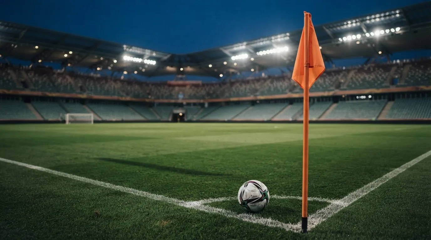Ein Fußball liegt neben einem Eckfahnen-Pfosten auf einem Rasenplatz, im Hintergrund ein leeres Stadion in der Dämmerung.
