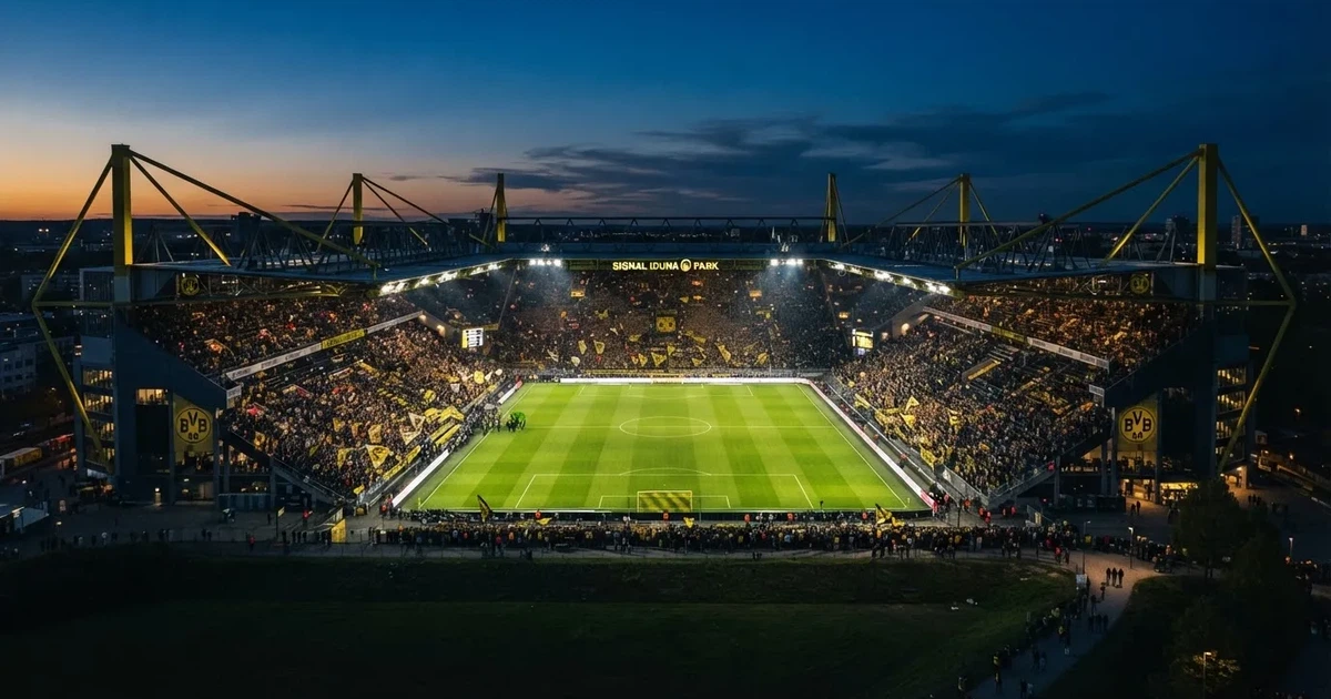Ein großes Stadion bei Nacht, aus der Ferne unter Flutlicht gesehen, die Ränge hell erleuchtet, der Rasen leuchtend grün.