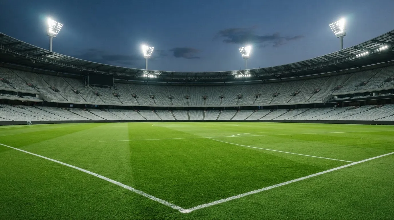 Eine leere Stadiontribüne unter Flutlicht aus der Ferne betrachtet, die Rasenfläche leuchtet grün in der Dämmerung.