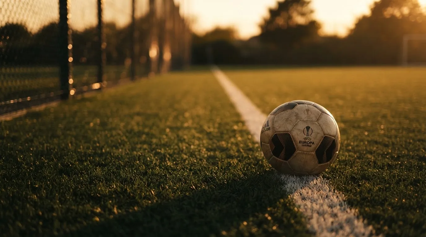 Ein Fußball liegt am Rand eines Rasens neben einer weißen Linie, im Hintergrund ein Zaun in der Abendsonne.