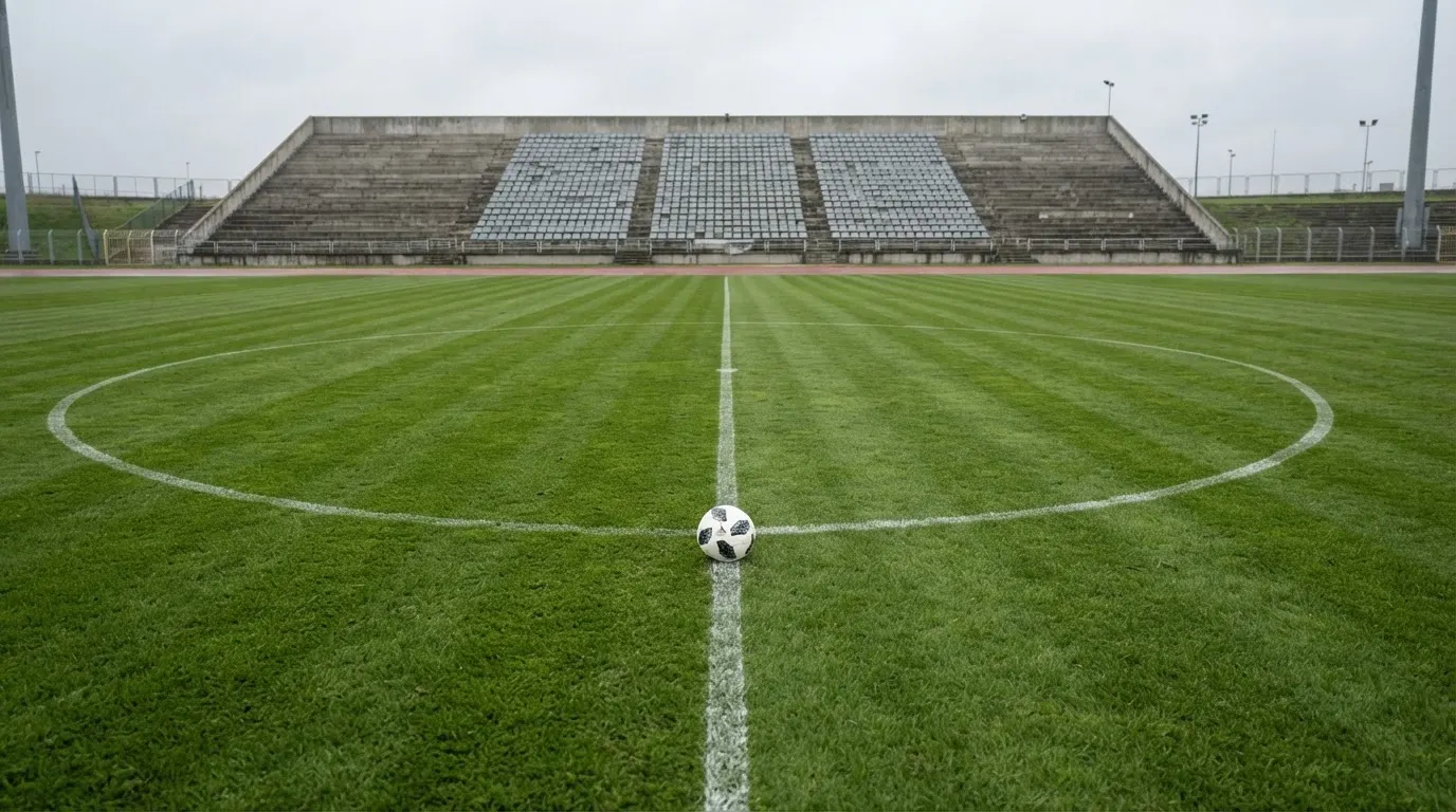 Ein Fußball liegt auf dem Anstoßpunkt eines leeren Rasenplatzes, im Hintergrund eine ruhige Tribüne unter bedecktem Himmel.