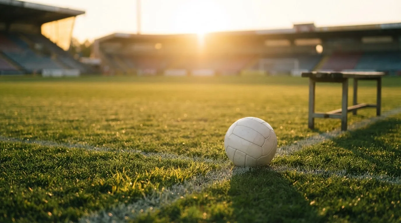 Ein Fußball auf einem Rasenplatz im Gegenlicht der tiefstehenden Sonne, am Spielfeldrand eine leere Ersatzbank.