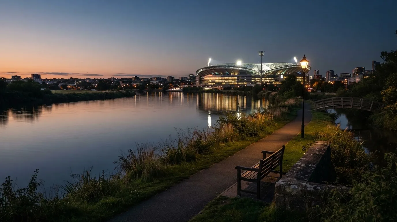 Ein Stadion unter Flutlicht aus der Ferne gesehen, im Vordergrund ein ruhiger Fluss und ein Fußweg.