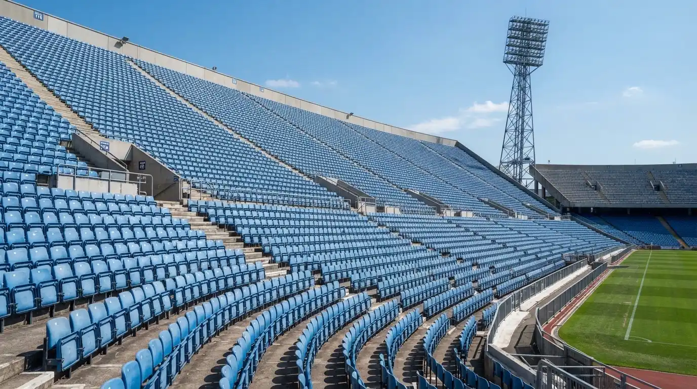 Eine leere Tribüne eines Fußballstadions bei Tageslicht, Sitzschalen in Reihen und dahinter ein Flutlichtmast.