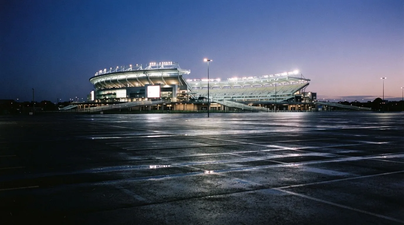 Ein Stadion unter Flutlicht aus der Ferne gesehen, davor ein leerer Parkplatz in der Dämmerung.