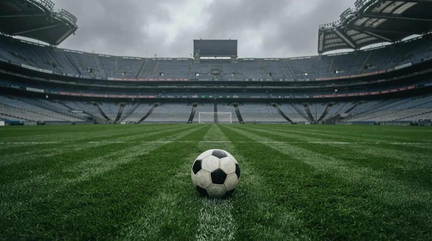 Ein Fußball auf einem Rasen, im Hintergrund ein leeres Stadion bei bewölktem Himmel.