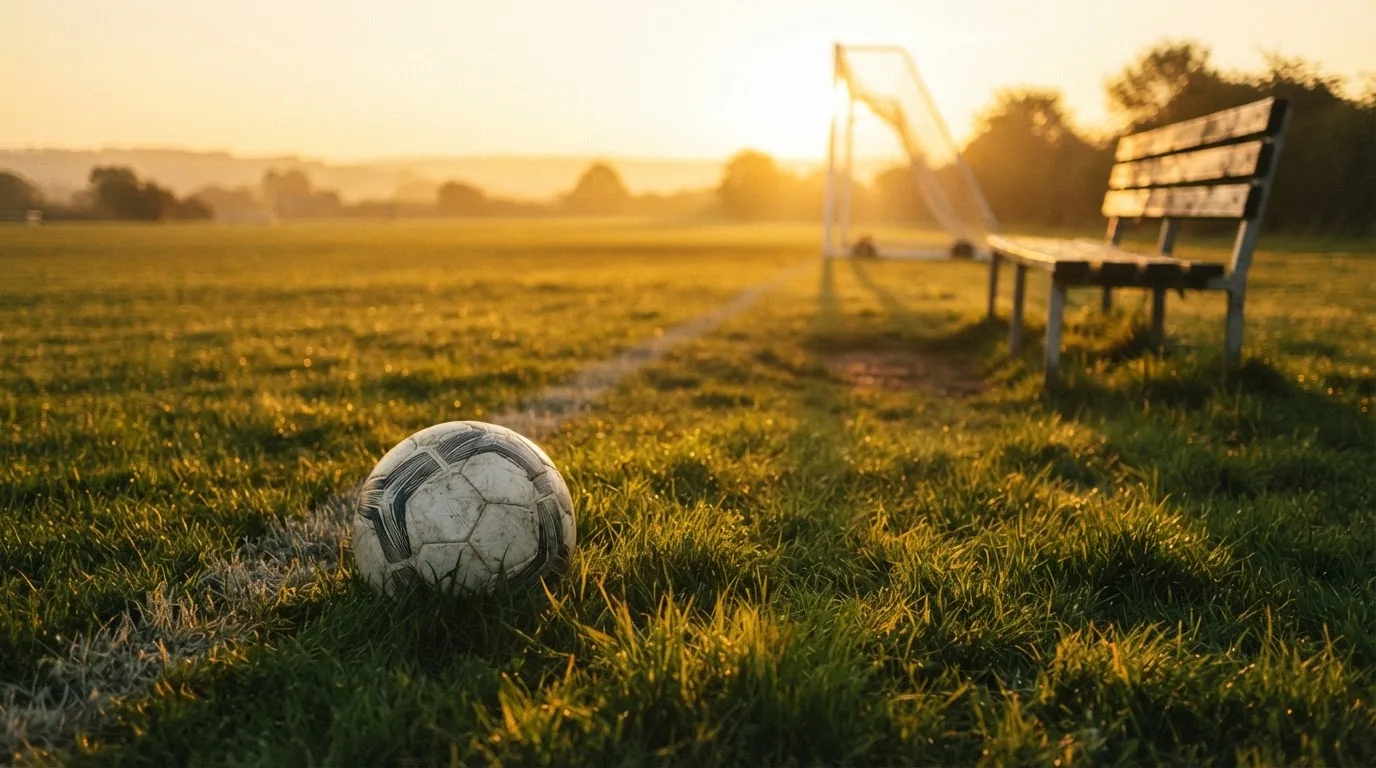 Ein Fußball liegt im Gras am Spielfeldrand, dahinter eine leere Bank und ein Tor im Gegenlicht.