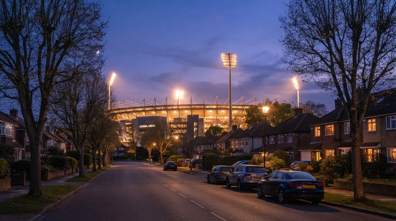 Ein Stadion aus der Ferne bei Flutlicht, davor eine ruhige Straße in der Abenddämmerung.