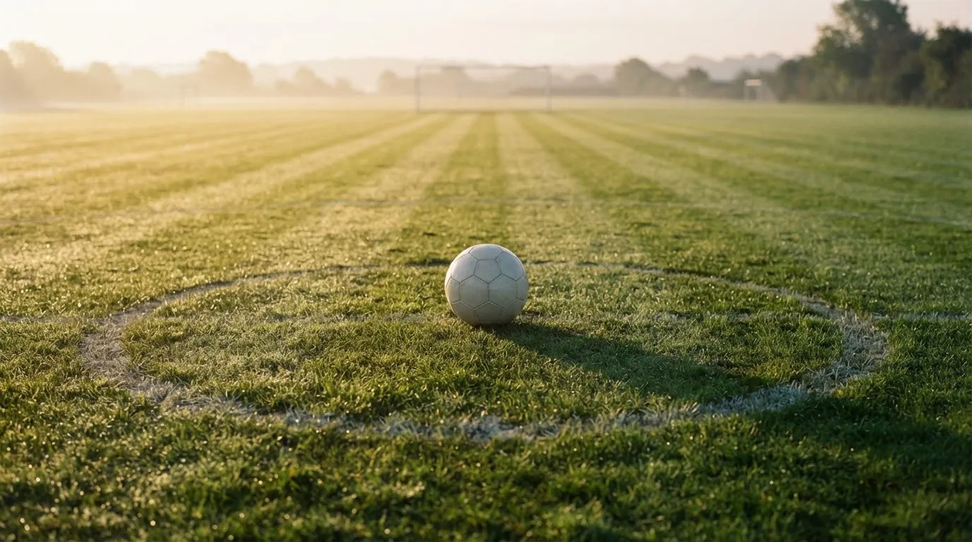 Ein Fußball auf dem Mittelkreis eines leeren Rasenplatzes im frühen Morgenlicht.