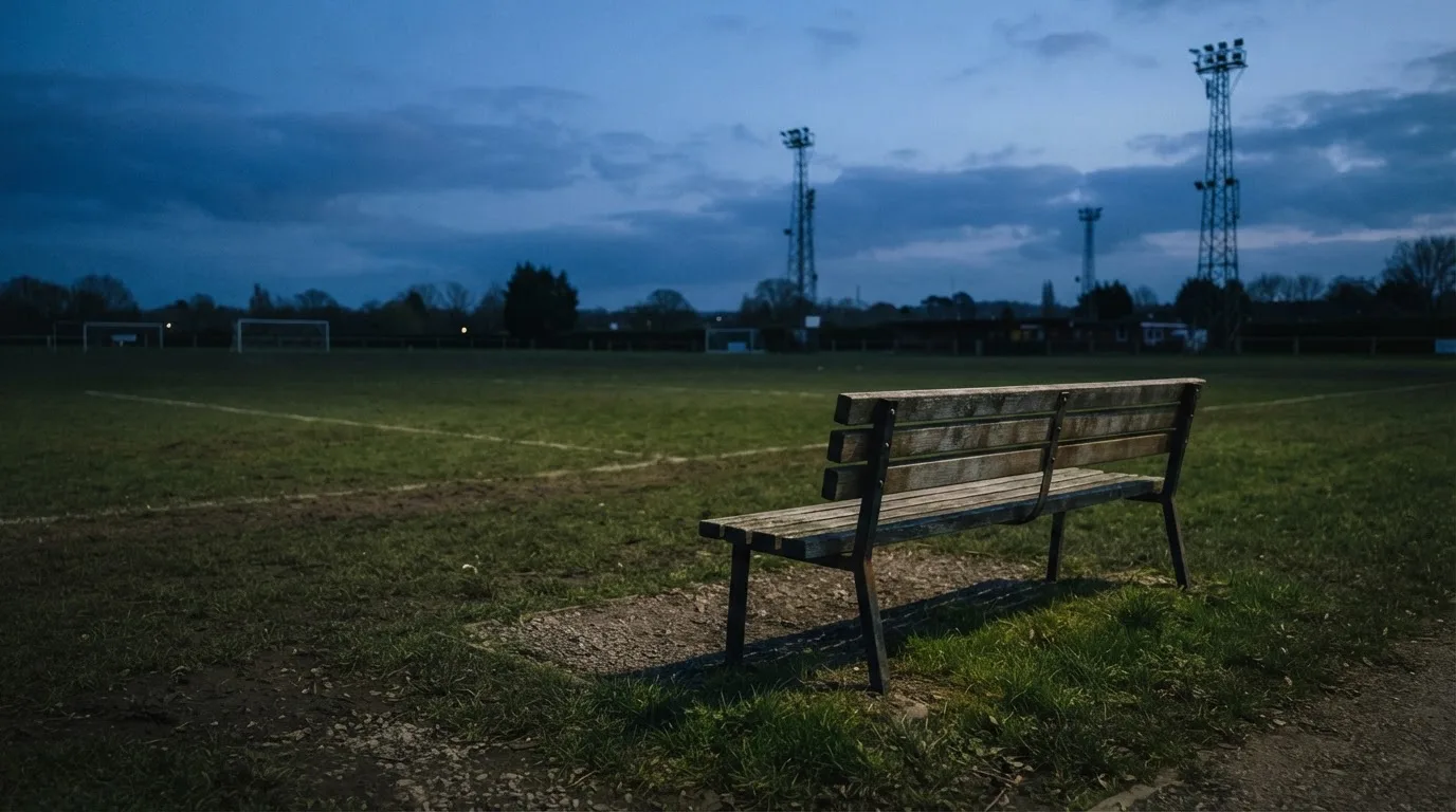 Eine leere Parkbank am Rand eines Fußballfelds in der Abenddämmerung, im Hintergrund ragen dunkle Flutlichtmasten in den Himmel.
