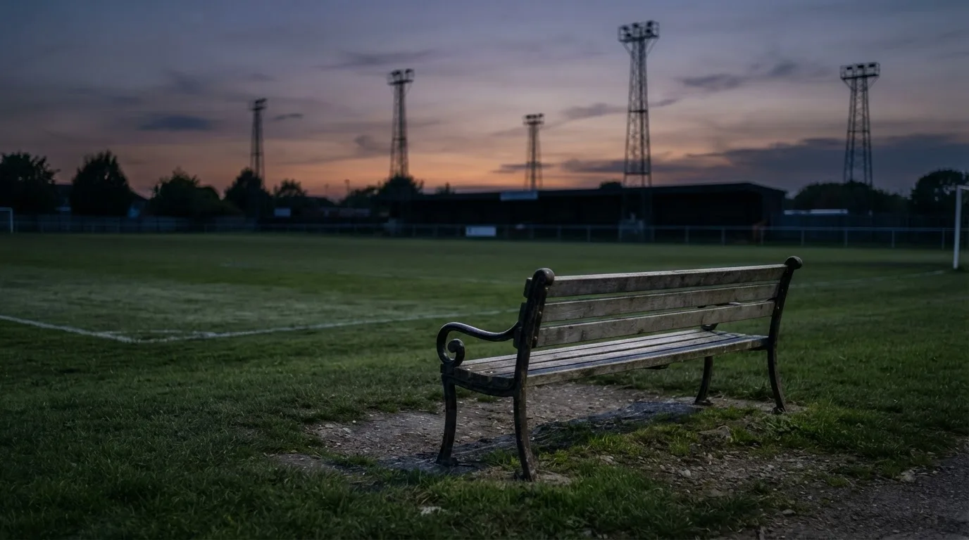 Eine leere Parkbank am Rand eines Fußballfelds in der Abenddämmerung, im Hintergrund ragen dunkle Flutlichtmasten in den Himmel.