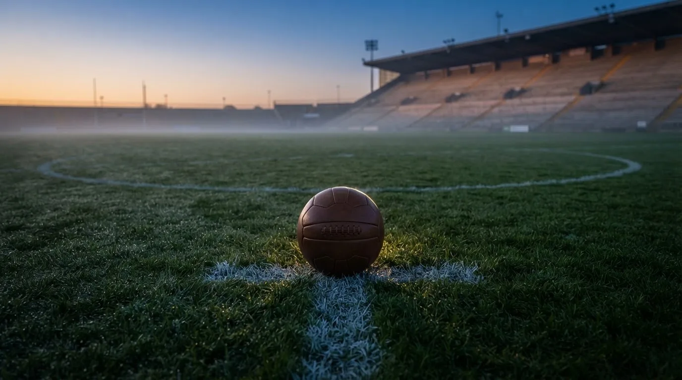 Ein Fußball liegt auf dem Anstoßpunkt eines leeren Rasenplatzes in der Morgendämmerung, im Hintergrund eine leere Tribüne.