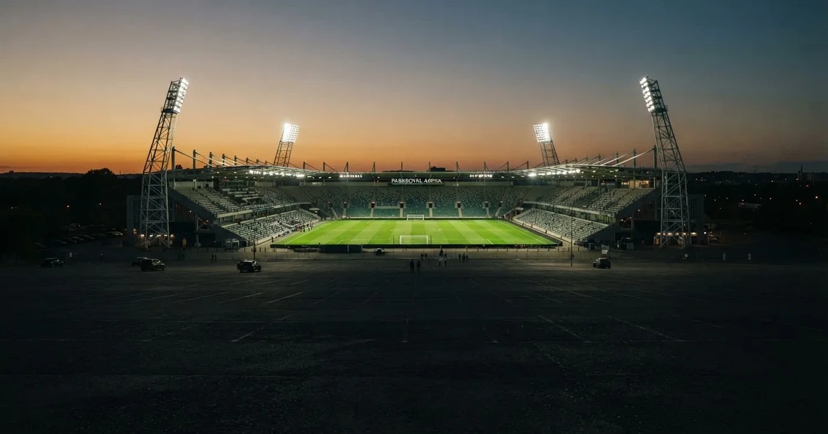 Ein Fußballstadion unter Flutlicht aus großer Entfernung, davor ein dunkler Parkplatz und ein heller Abendhimmel.