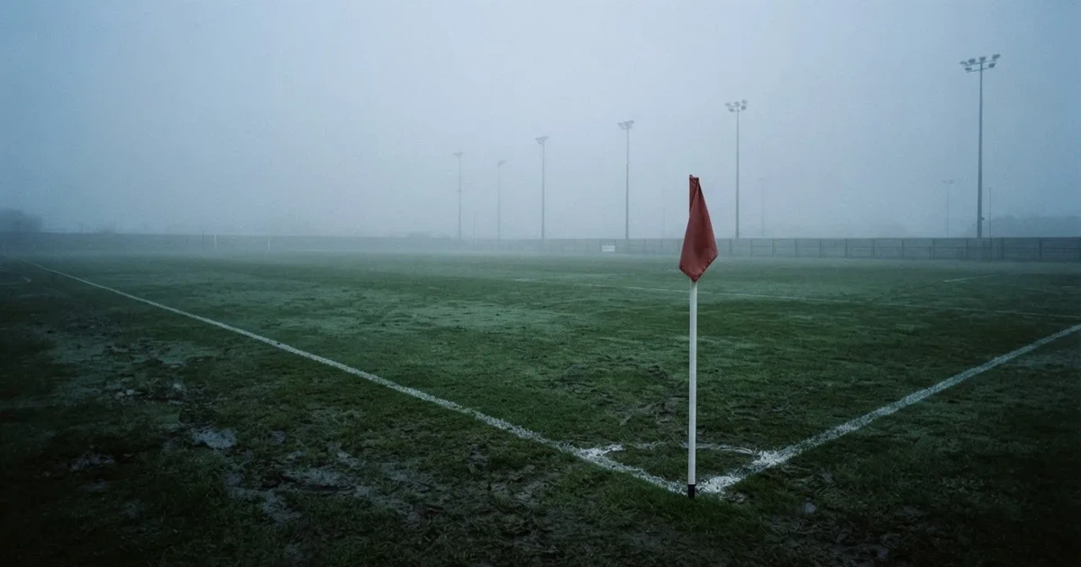 Eine Eckfahne steht im Nebel am Rand eines leeren Rasenplatzes in der Morgendämmerung, dahinter ein Flutlichtmast.