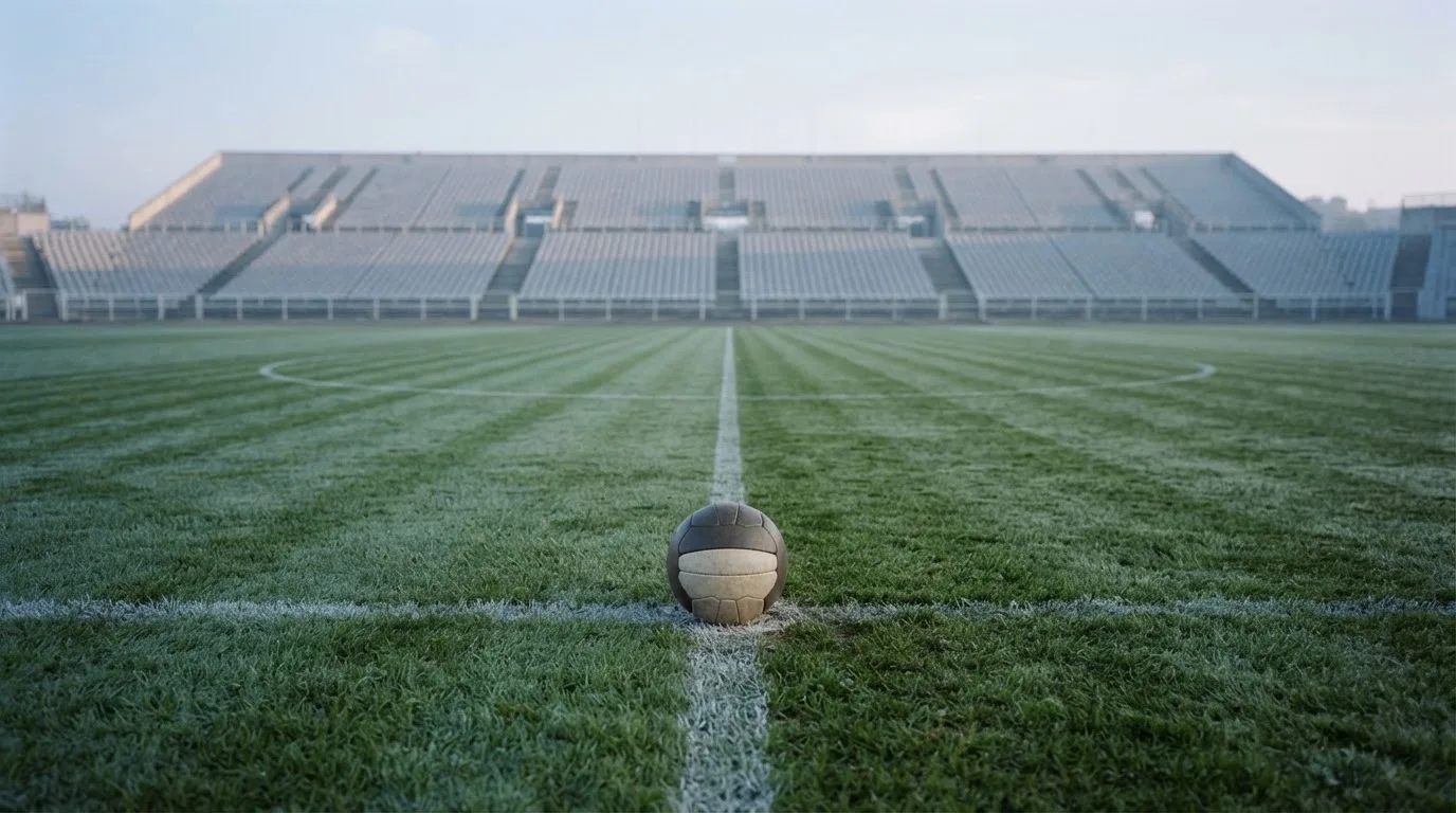 Ein Fußball liegt auf dem Anstoßpunkt eines leeren Rasenplatzes im frühen Morgenlicht, dahinter eine leere Tribüne.