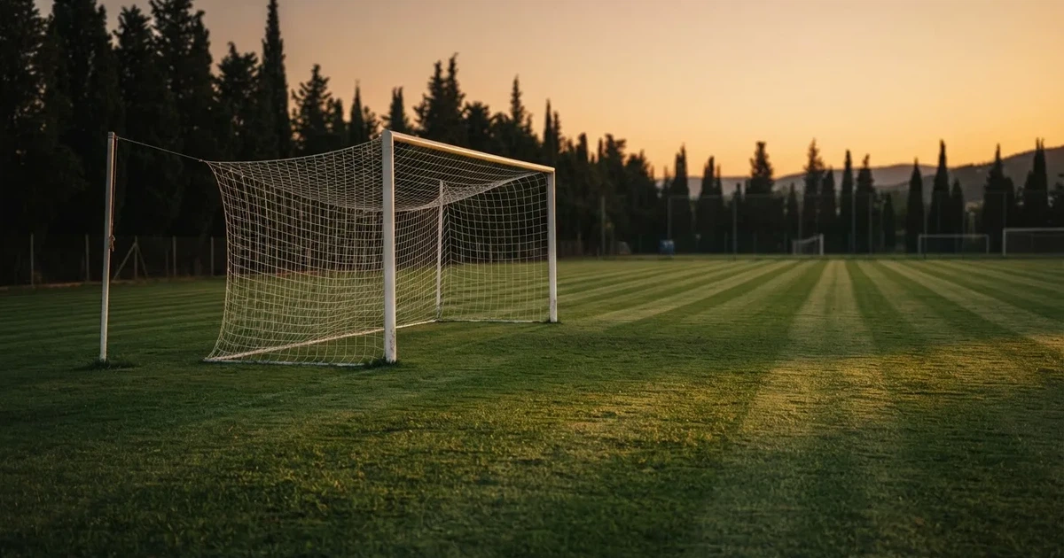 Ein Fußballtor mit Netz auf einem leeren Platz im Abendlicht, der Rasen frisch gemäht, im Hintergrund Zypressen.