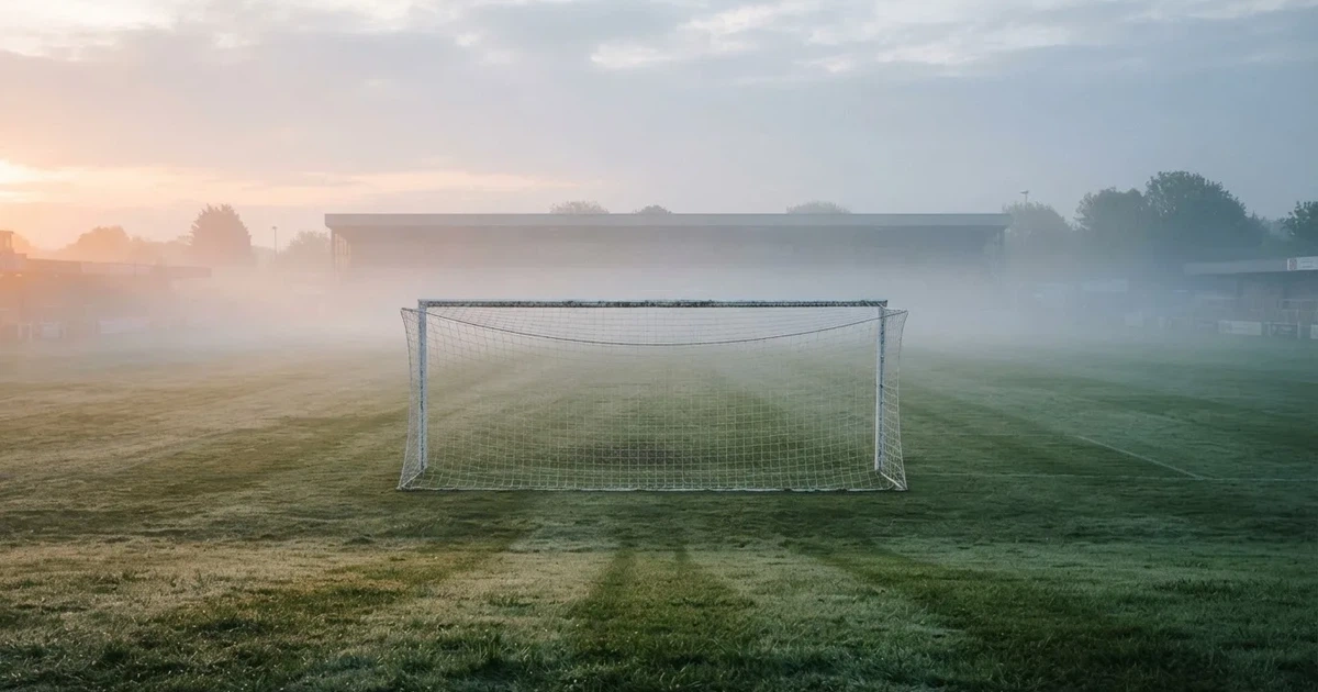 Ein Fußballtor mit Netz steht auf einem leeren Platz im Nebel der Morgendämmerung, der Rasen ist taufeucht.