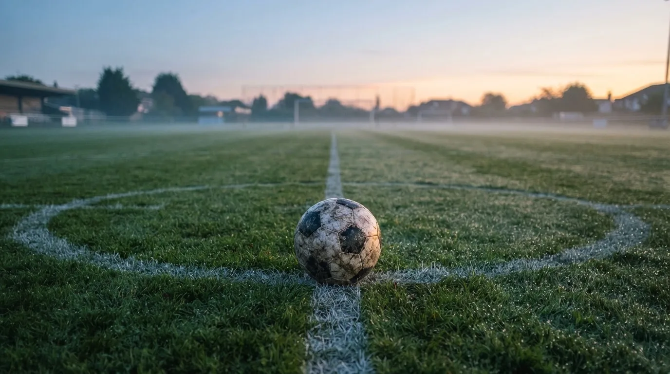Ein Fußball ruht auf dem Mittelkreis eines leeren Rasenplatzes in der Morgendämmerung, Tau liegt auf dem Gras.
