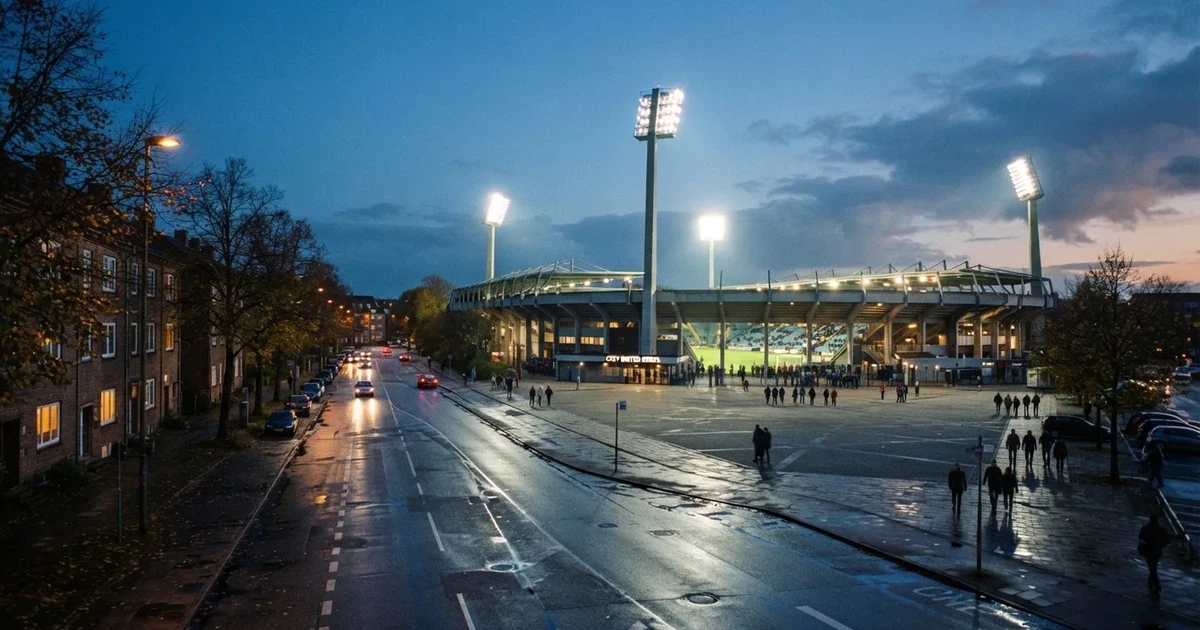 Ein Fußballstadion bei Flutlicht aus der Ferne gesehen, davor eine Straße und leuchtende Fenster eines Nebengebäudes.