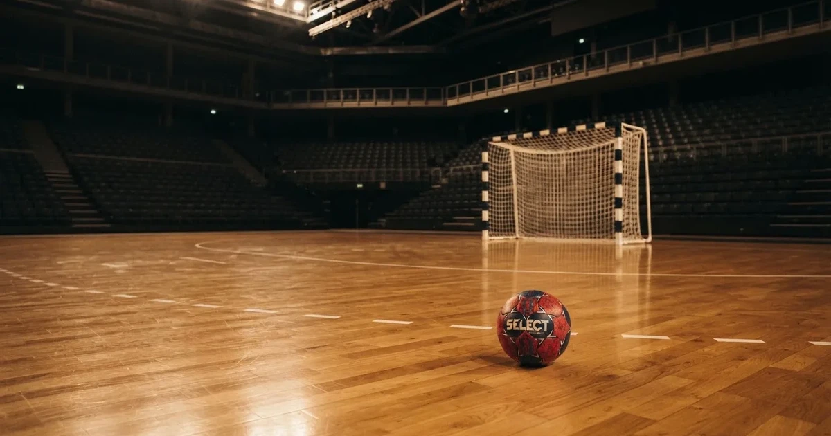 Ein Handball liegt auf dem Hallenboden vor einem Tor mit Netz, die leere Halle wird von Flutlicht beleuchtet.