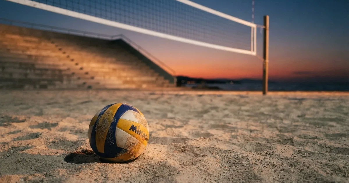 Ein Volleyball liegt auf dem Sand eines Beachvolleyball-Feldes neben dem gespannten Netz, im Hintergrund eine leere Tribüne bei Abendlicht.