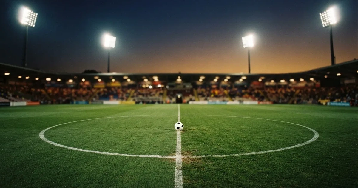 Ein Rasenplatz in der Dämmerung mit eingeschalteten Flutlichtmasten, ein Ball liegt am Anstoßpunkt, die Tribünen im Hintergrund sind unscharf.