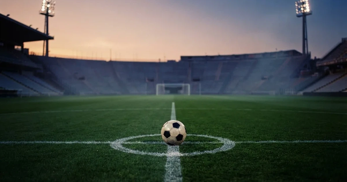Ein Fußball liegt auf dem Anstoßpunkt eines leeren Rasenplatzes, im Hintergrund eine Tribüne und Flutlichtmasten in der Abenddämmerung.