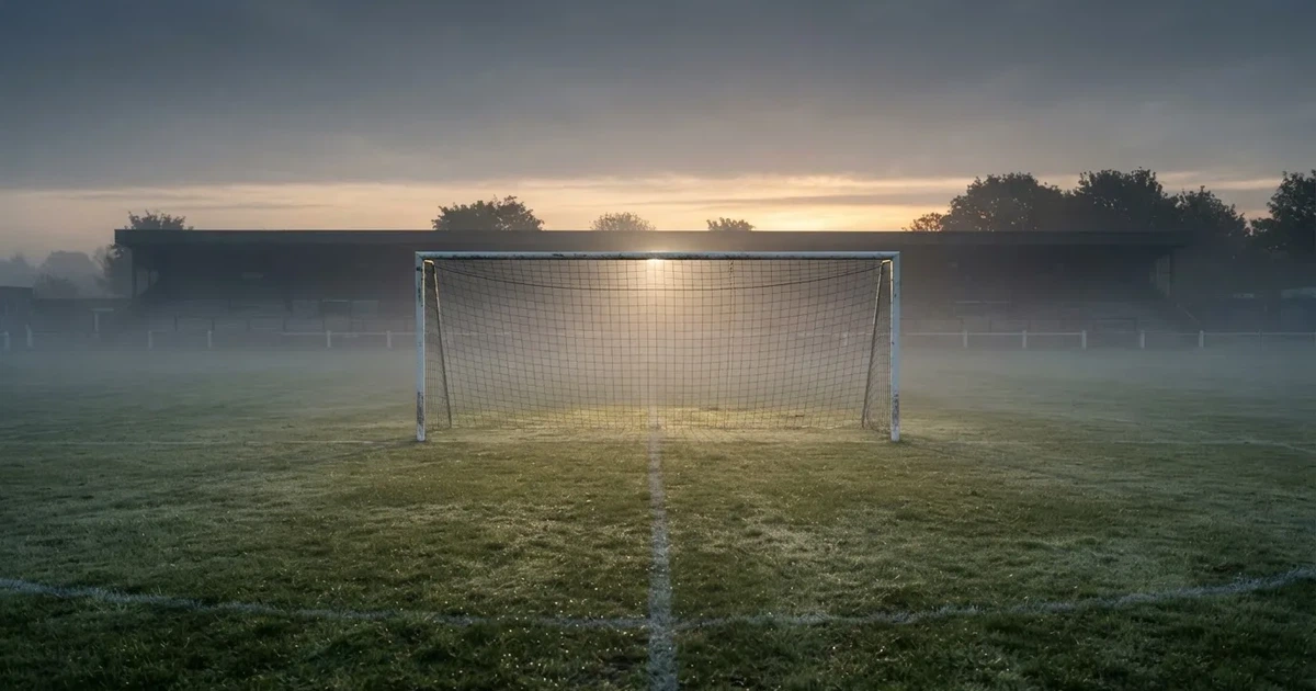 Ein Fußballtor mit Netz steht im Morgennebel auf einem leeren Rasenplatz, im Hintergrund die Silhouette einer kleinen Tribüne.