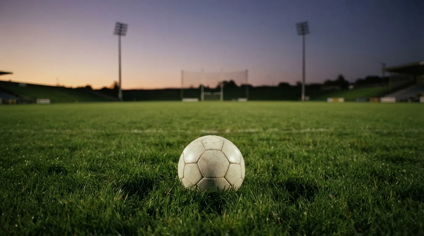 Ein Fußball liegt im Gras eines leeren Platzes, im Hintergrund stehen Flutlichtmasten in der Dämmerung.
