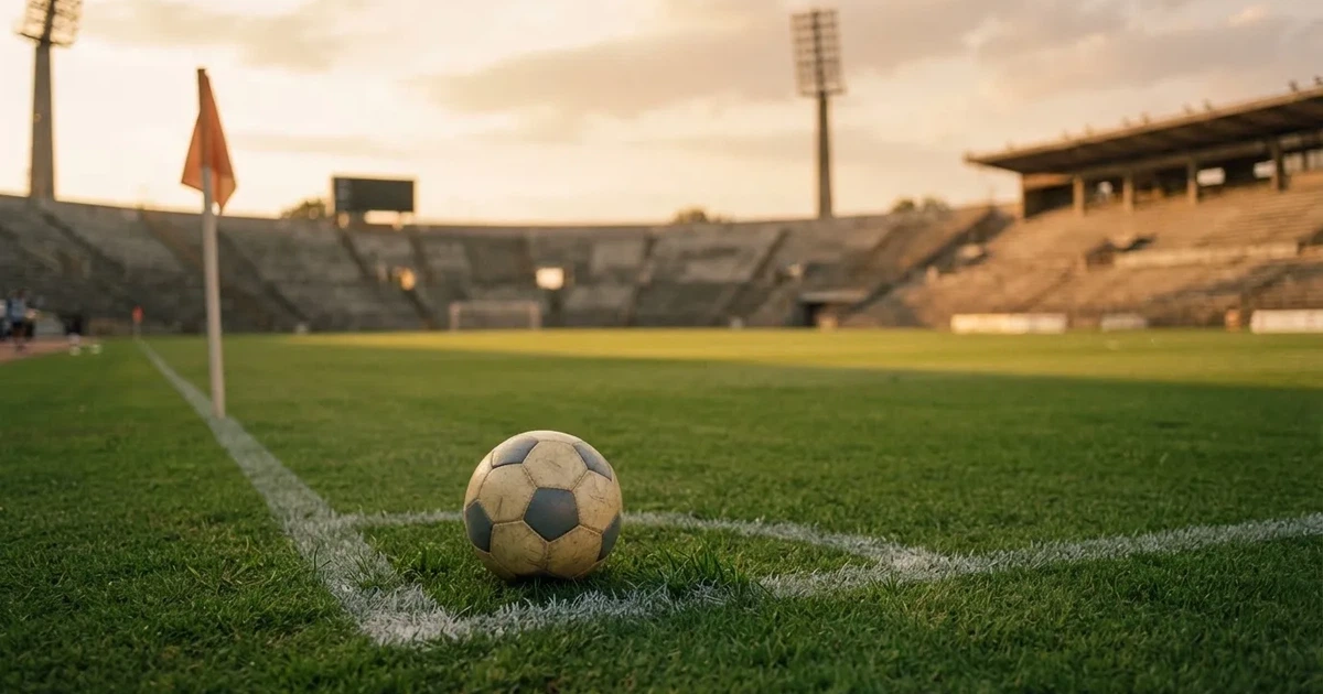 Ein Fußball liegt am Viertelkreis an der Eckfahne eines Rasenplatzes, im Hintergrund leere Tribünen in warmem Abendlicht.