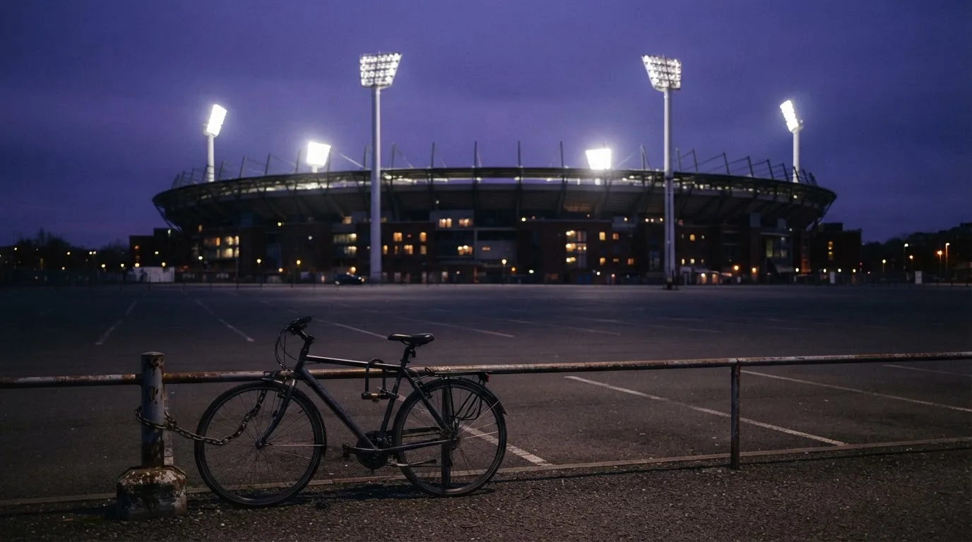 Ein Stadion unter Flutlicht aus der Ferne gesehen, davor ein dunkler Parkplatz und ein einzelnes Fahrrad an einem Zaun.