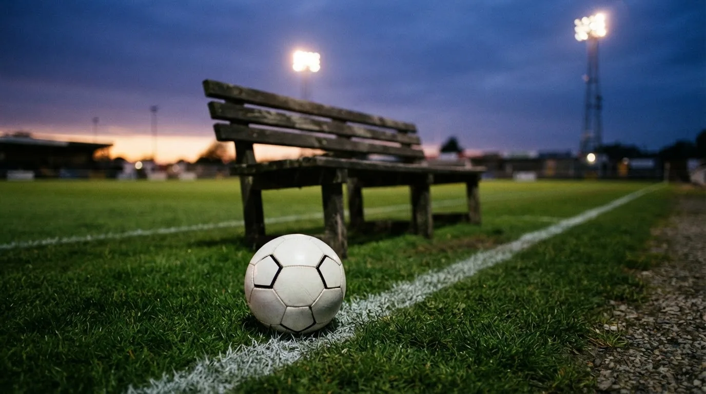 Ein Fußball liegt am Spielfeldrand neben einer leeren Ersatzbank, das Flutlicht über dem Platz leuchtet in der Dämmerung.