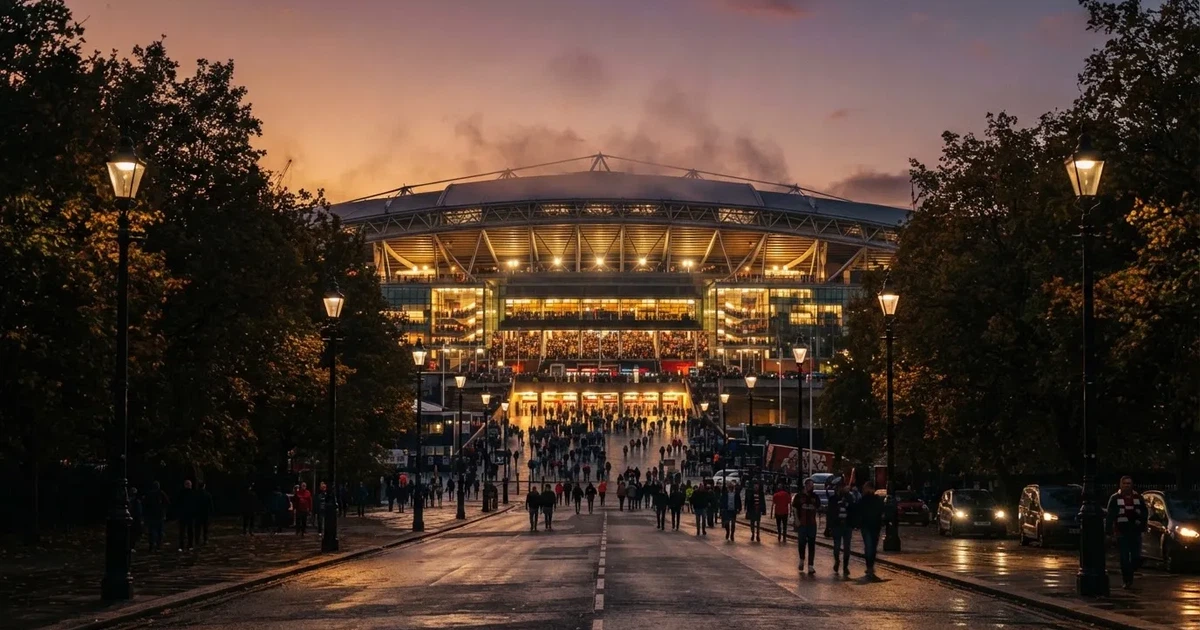 Ein großes Fußballstadion mit hell erleuchtetem Rund, aus der Ferne am Abend gesehen, davor eine breite Allee mit Laternen.