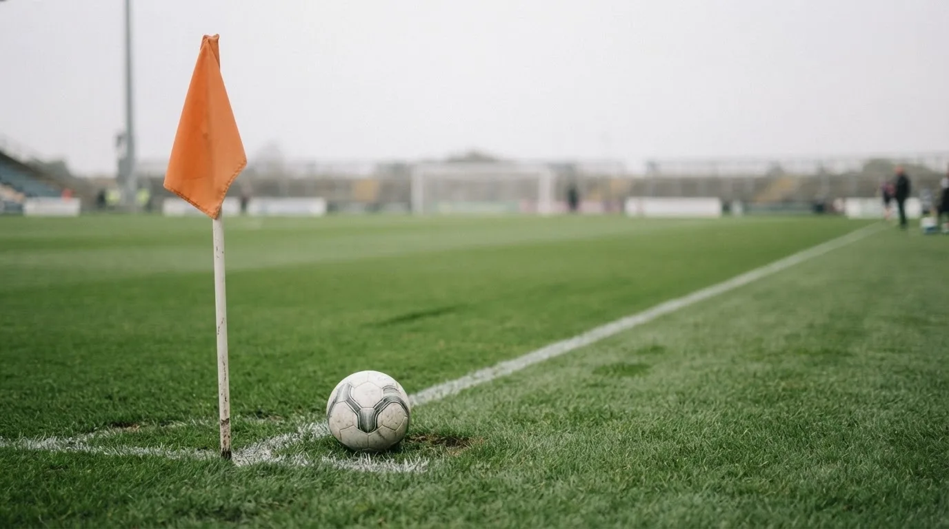 Ein Fußball liegt neben einer Eckfahne auf kurz gemähtem Rasen unter bewölktem Himmel.